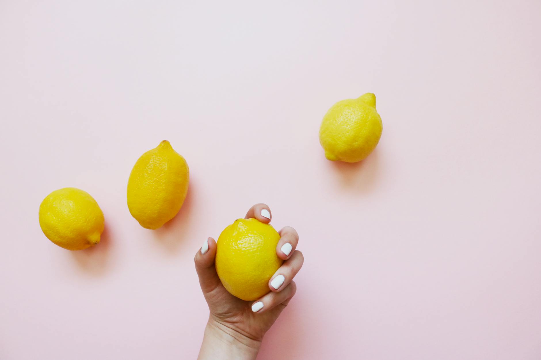 Una mano sosteniendo un limón amarillo brillante sobre un fondo rosa suave, rodeado de limones adicionales