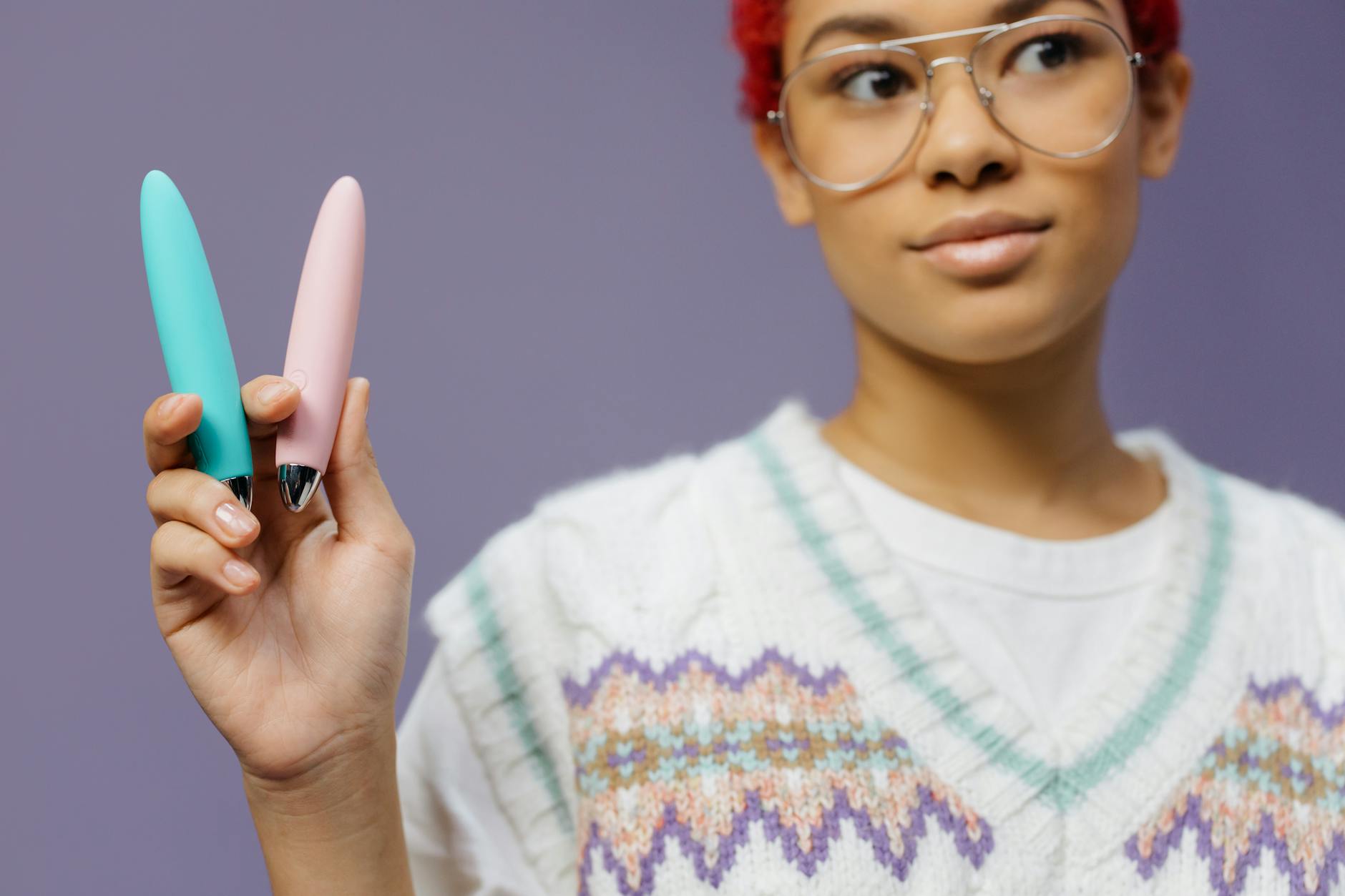 A person holds two different clitoral vibrators side by side against a purple background.